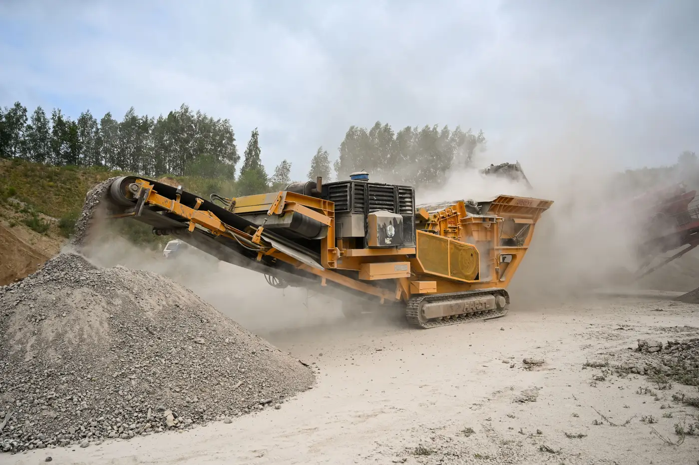 A yellow tracked mobile crusher churning rock and dust next to a gravel stockpile, with trees and an overcast sky behind it.